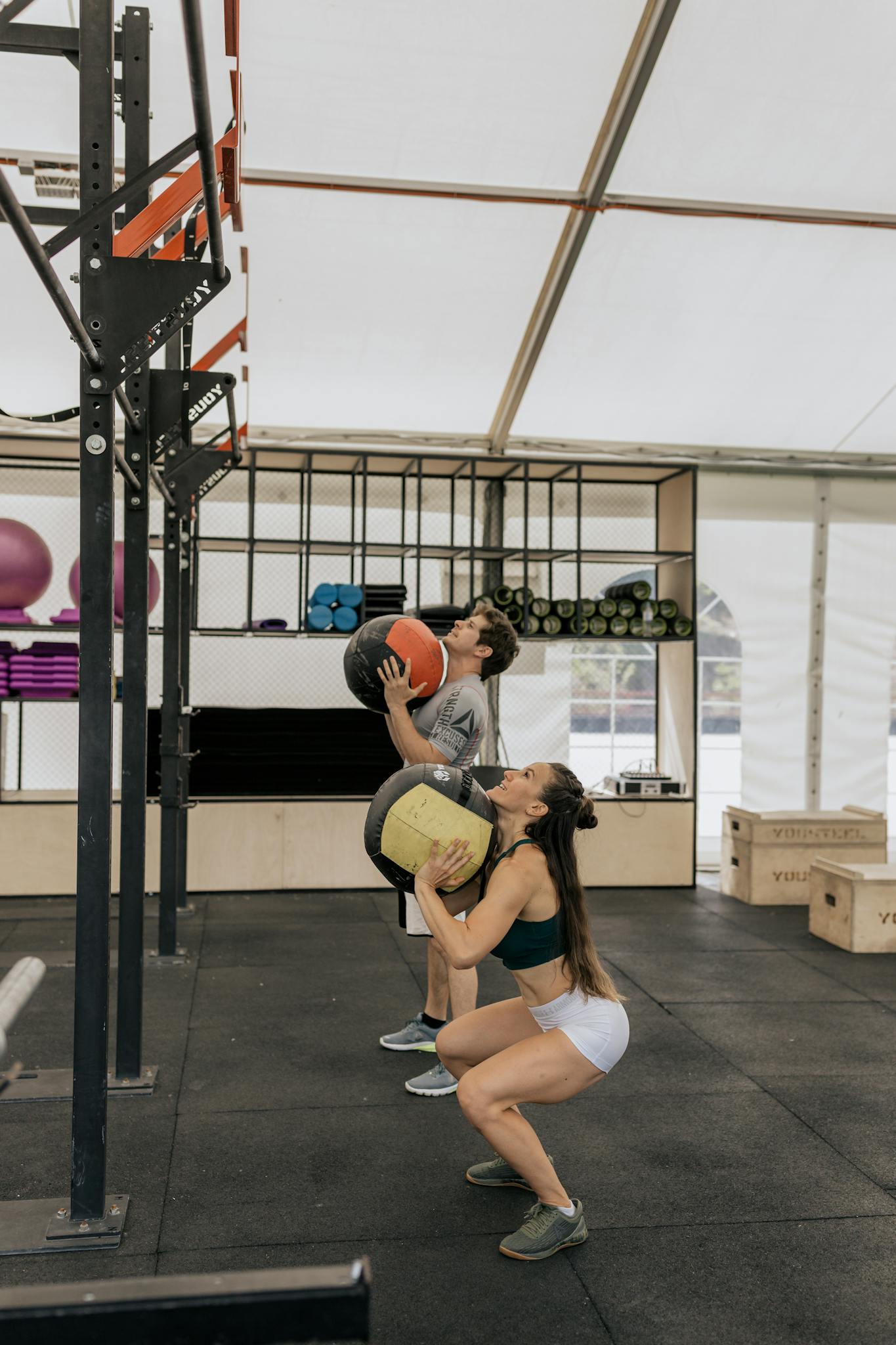 A man and woman working out with medicine balls in an indoor gym setting.